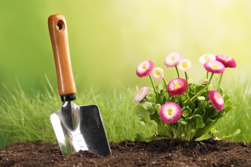 In the image, a small gardening scene features a gardening trowel with a wooden handle and a metal scoop partially inserted into rich, dark soil, indicating recent planting activity. To the right, there is a vibrant cluster of pink and white primula flowers with round, full blooms and green foliage, adding colour and natural beauty. The background showcases a soft focus, lush green grassy area, suggesting an outdoor garden setting in good weather conditions. The soil appears well-maintained and freshly turned, suitable for planting, while the grass is evenly cut and healthy. This scene emphasizes typical gardening work and outdoor landscape features relevant to services such as garden planting, soil preparation, and flower bed maintenance, aligning with the services provided by Gardening Services Bethnal Green. The natural tones of the soil and greenery contrast with the bright blooms, creating a visually appealing and informative image that supports gardening and landscaping themes in a residential and urban context in Bethnal Green, London, within the E2 postcode area.