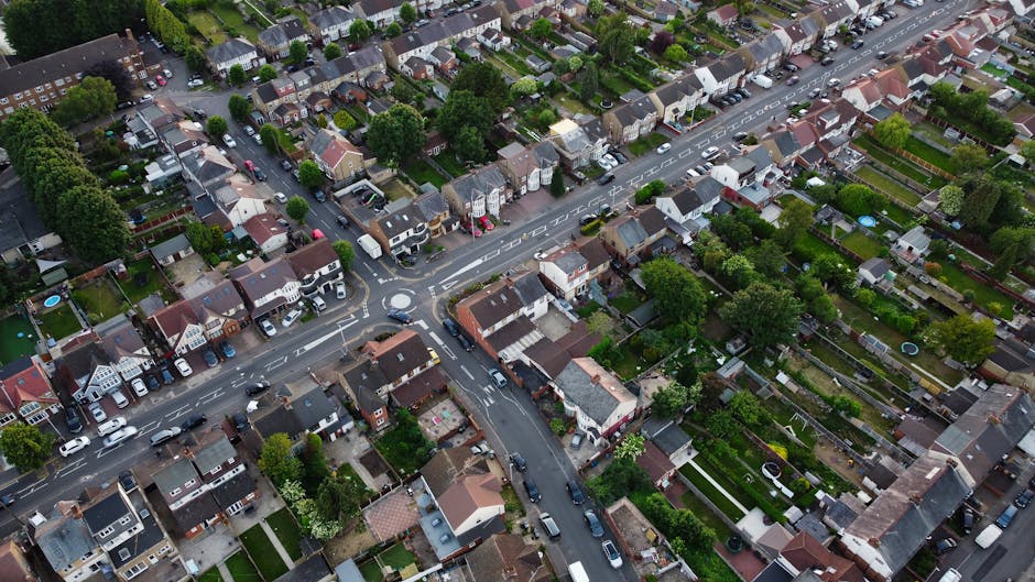 An aerial view of a residential neighborhood featuring a mix of detached and terraced houses with front gardens and backyards. The scene includes well-maintained lawns, hedges, and garden beds with va